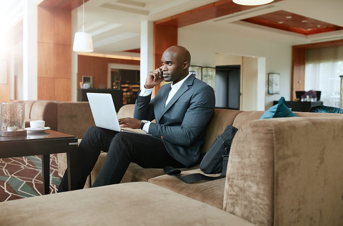 Businessman sitting on sofa with a laptop using mobile phone. Busy african male executive waiting in hotel lobby.