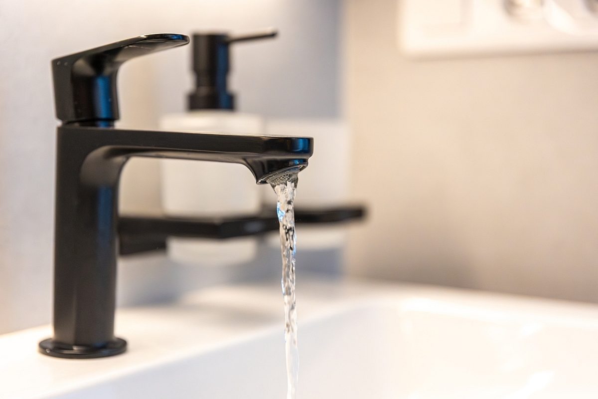 Matte black faucet pours a steady stream over a white sink with a soft bokeh soap dispenser behind. Clean lines and minimalist style for a contemporary interior.