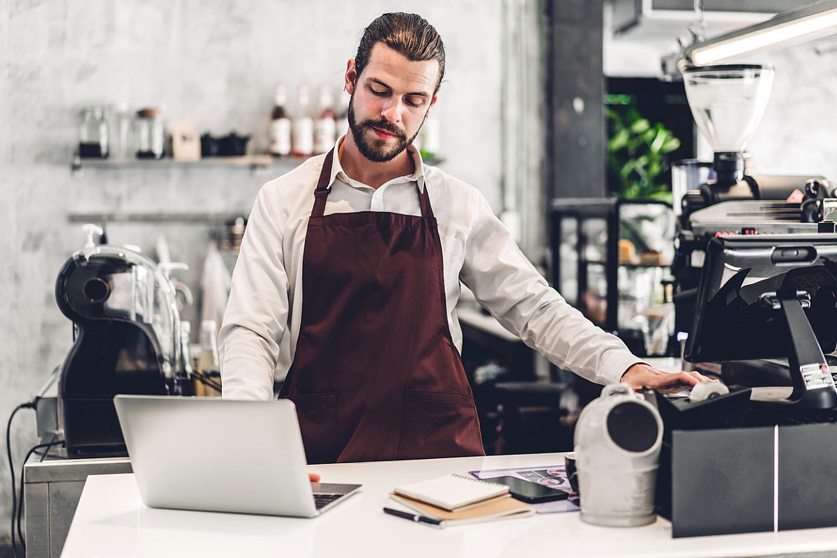 Portrait of handsome bearded barista man small business owner working with laptop computer behind the counter bar in a cafe