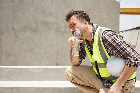 Dangerous from heat wave during working outside the building. Caucasian male construction worker resting and drinking water from bottle in a very hot day. Heat stroke health problem.