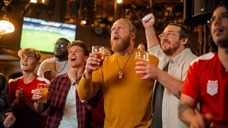 Group of Friends Watching a Live Soccer Match on TV in a Sports Bar. Three Men Cheering and Shouting. Young People Celebrating and Toasting Beer Glasses When Team Wins the Football World Cup.